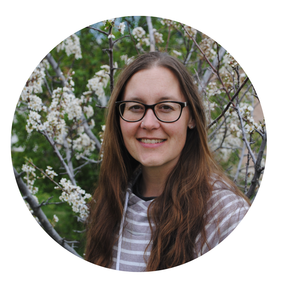 A woman with long hair and glasses smiles standing in front of a tree filled with spring blossoms.
